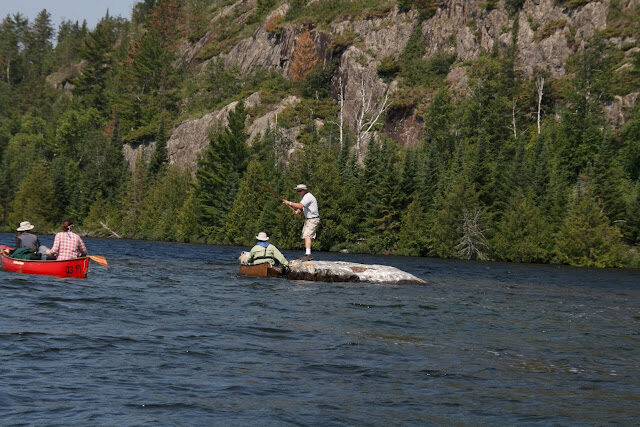 An angler casting below a rugged cliff face while a red canoe waits nearby